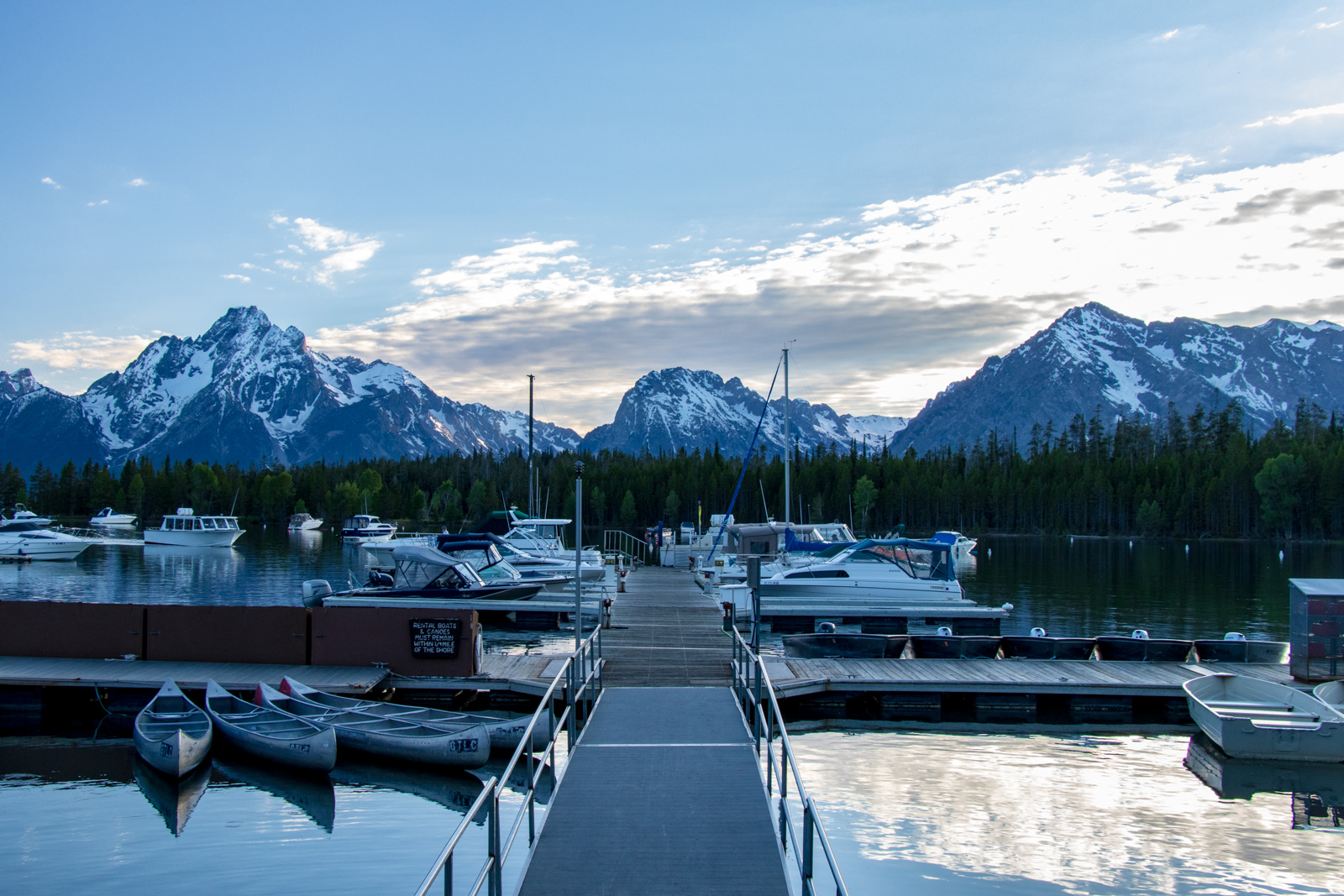Boats docked at a mountain marina with the Teton Range reflected in calm water at dusk
